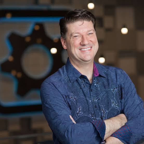 Randy Pitchford smiling with his arms locked, wearing a a full-sevee shirt